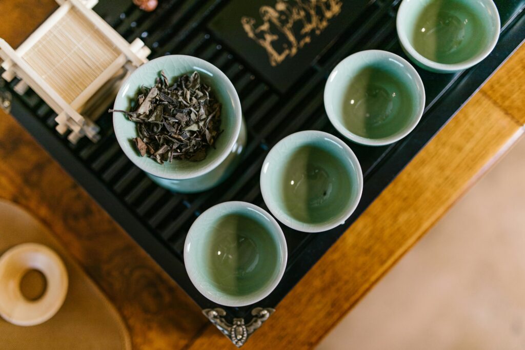 Top view of a traditional tea set with green teacups and dried tea leaves on a bamboo rack, captured in a warm setting.